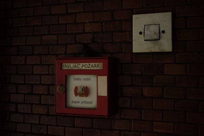 A red fire alarm box is mounted on a dark brick wall. The box has a label with text written in a foreign language, possibly indicating instructions or information for emergency situations. Next to the box is a square, white electrical switch panel.