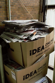 Two large cardboard boxes are stacked on top of each other, filled with a pile of newspapers and magazines. The boxes are labeled with 'IDEA ONLINE PRODAVNICA.' The setting appears industrial or storage-like, with brick walls and metal pipes visible in the background.