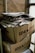 Two large cardboard boxes are stacked on top of each other, filled with a pile of newspapers and magazines. The boxes are labeled with 'IDEA ONLINE PRODAVNICA.' The setting appears industrial or storage-like, with brick walls and metal pipes visible in the background.