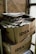 Two large cardboard boxes are stacked on top of each other, filled with a pile of newspapers and magazines. The boxes are labeled with 'IDEA ONLINE PRODAVNICA.' The setting appears industrial or storage-like, with brick walls and metal pipes visible in the background.