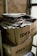 Two large cardboard boxes are stacked on top of each other, filled with a pile of newspapers and magazines. The boxes are labeled with 'IDEA ONLINE PRODAVNICA.' The setting appears industrial or storage-like, with brick walls and metal pipes visible in the background.