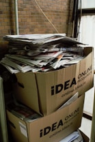 Two large cardboard boxes are stacked on top of each other, filled with a pile of newspapers and magazines. The boxes are labeled with 'IDEA ONLINE PRODAVNICA.' The setting appears industrial or storage-like, with brick walls and metal pipes visible in the background.