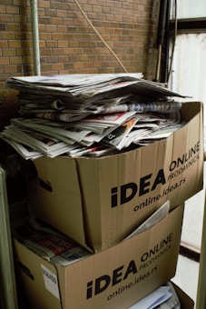Two large cardboard boxes are stacked on top of each other, filled with a pile of newspapers and magazines. The boxes are labeled with 'IDEA ONLINE PRODAVNICA.' The setting appears industrial or storage-like, with brick walls and metal pipes visible in the background.