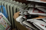 A large stack of newspapers is piled in an open cardboard box next to an old radiator. Nearby, a bundle of newspapers is wrapped in a blue plastic bag. The newspapers contain colorful images and headlines.