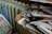 A large stack of newspapers is piled in an open cardboard box next to an old radiator. Nearby, a bundle of newspapers is wrapped in a blue plastic bag. The newspapers contain colorful images and headlines.