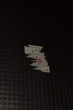 A partially peeled sticker is stuck on a dark tiled surface. The sticker has blue and red stars and some incomplete text visible.