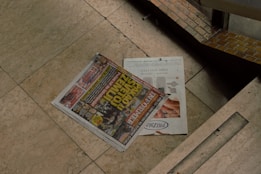 A newspaper with bold headlines and colorful images is placed on a light-colored marble floor. The newspaper features advertisements and various news stories. Adjacent to it is a magazine with an advertisement displayed, partially covered by the newspaper. The surrounding area appears to be part of an architecture with tiles and a darker shadowy corner.