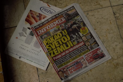 A newspaper is placed on a tiled floor. The headline in large yellow letters stands out. Next to it, there is an advertisement showing electronic massage devices. The overall setting seems casual and informal.