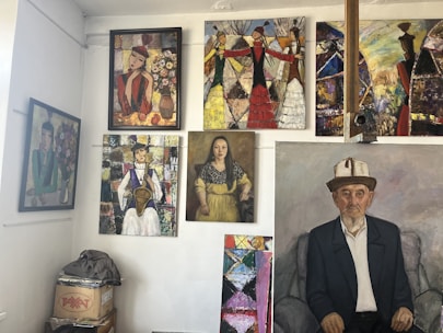 A gallery wall featuring multiple paintings. The artworks include various portraits and abstract pieces, showcasing people in traditional attire and colorful abstract patterns. The floor has a stack of boxes with objects on top.