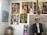 A gallery wall featuring multiple paintings. The artworks include various portraits and abstract pieces, showcasing people in traditional attire and colorful abstract patterns. The floor has a stack of boxes with objects on top.
