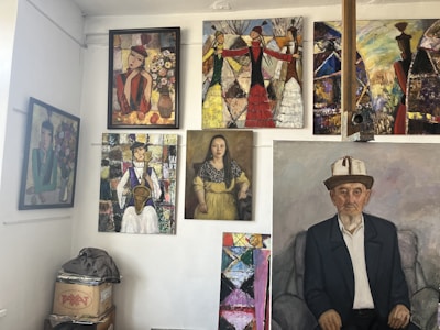 A gallery wall featuring multiple paintings. The artworks include various portraits and abstract pieces, showcasing people in traditional attire and colorful abstract patterns. The floor has a stack of boxes with objects on top.