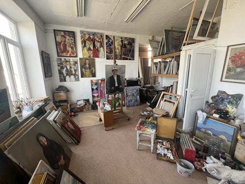 An art studio filled with paintings, canvases, and art supplies. Various completed and in-progress artworks are displayed on the walls and stacked around the room. A central easel holds a portrait, and surrounding it are brushes, palettes, and other tools. Shelves hold frames and more paintings, while natural light streams in through a large window.