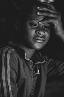 A person is depicted in close-up with a somber expression, showing a tear on their cheek. Their hand rests on their forehead and they are wearing a zip-up jacket with a stripe on the sleeve.