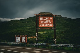 A rustic wooden sign with white letters welcomes visitors to 'Colorful Colorado' against a backdrop of lush, green mountains. The sky is overcast with dark clouds, suggesting an impending storm. Additional signs are visible off to the side, surrounded by a guardrail covered in graffiti.
