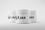 Three white jars with ribbed lids are arranged on a light background. The jars are labeled with the text 'JAR' accompanied by a minimalist graphic logo. They appear to be mockup templates for product design presentation.