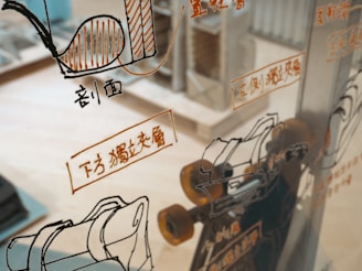 A glass surface reflects various technical schematics and illustrations, likely related to machinery or engineering, with accompanying text in a foreign language. The background shows a blurred interior space, possibly a workshop or an office.
