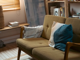 A cozy living room featuring a mid-century modern style sofa with olive green upholstery, adorned with a decorative pillow in shades of blue and cream. The room is well-lit with natural light filtering through wooden blinds. A wooden shelf is visible in the background, filled with books and decor. A woven basket sits next to a small table with a stack of books.