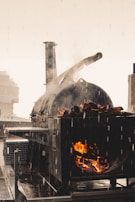 A large outdoor cooking smoker is in use, with visible flames and smoke rising from it. The surrounding environment suggests a rainy day, as droplets of water can be seen. The smoker is loaded with logs, and the flames create a warm contrast against the gray and rainy atmosphere.