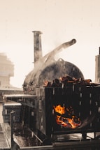 A large outdoor cooking smoker is in use, with visible flames and smoke rising from it. The surrounding environment suggests a rainy day, as droplets of water can be seen. The smoker is loaded with logs, and the flames create a warm contrast against the gray and rainy atmosphere.