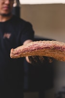 A person holds a seasoned piece of meat, likely ribs, with a visible crust of spices and seasonings. The focus is on the meat, which appears to be raw, showcasing a marbled texture.