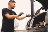 A person wearing a black t-shirt and gloves is tending to a large barbecue smoker, adjusting pieces of meat. The setting appears to be outdoors with a building visible in the background. The image conveys a sense of focus and culinary activity.