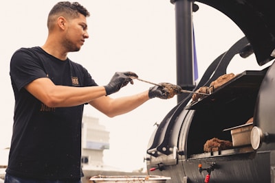 A person wearing a black t-shirt and gloves is tending to a large barbecue smoker, adjusting pieces of meat. The setting appears to be outdoors with a building visible in the background. The image conveys a sense of focus and culinary activity.