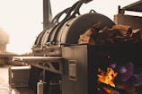 A large industrial smoker is positioned outdoors, with a fire burning inside to the side. Wood logs are stacked on top of the smoker, and sunlight bathes the area in a warm glow.