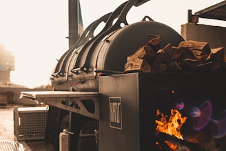 A large industrial smoker is positioned outdoors, with a fire burning inside to the side. Wood logs are stacked on top of the smoker, and sunlight bathes the area in a warm glow.