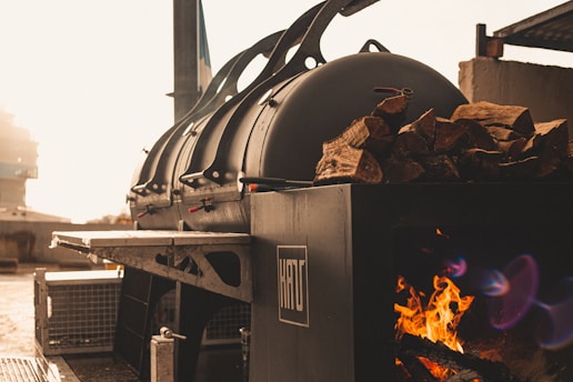 A large industrial smoker is positioned outdoors, with a fire burning inside to the side. Wood logs are stacked on top of the smoker, and sunlight bathes the area in a warm glow.