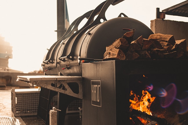 A large industrial smoker is positioned outdoors, with a fire burning inside to the side. Wood logs are stacked on top of the smoker, and sunlight bathes the area in a warm glow.