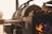 A large industrial smoker is positioned outdoors, with a fire burning inside to the side. Wood logs are stacked on top of the smoker, and sunlight bathes the area in a warm glow.