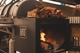 A large black industrial smoker is displayed, with a section open to reveal a burning fire inside. On top of the smoker is a pile of chopped firewood, ready to be used as fuel. The overall setting appears to be industrial or outdoor cooking-related, with metal elements surrounding the smoker.