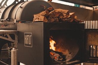 A large black industrial smoker is displayed, with a section open to reveal a burning fire inside. On top of the smoker is a pile of chopped firewood, ready to be used as fuel. The overall setting appears to be industrial or outdoor cooking-related, with metal elements surrounding the smoker.