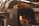 A large black industrial smoker is displayed, with a section open to reveal a burning fire inside. On top of the smoker is a pile of chopped firewood, ready to be used as fuel. The overall setting appears to be industrial or outdoor cooking-related, with metal elements surrounding the smoker.
