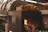 A large black industrial smoker is displayed, with a section open to reveal a burning fire inside. On top of the smoker is a pile of chopped firewood, ready to be used as fuel. The overall setting appears to be industrial or outdoor cooking-related, with metal elements surrounding the smoker.