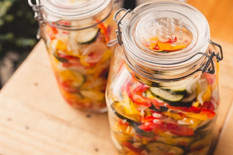 Two glass jars filled with colorful pickled vegetables, including slices of zucchini, red and yellow bell peppers, and onions. The jars have metal clasps and are placed on a light wooden surface.