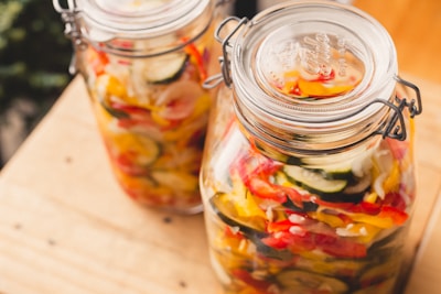 Two glass jars filled with colorful pickled vegetables, including slices of zucchini, red and yellow bell peppers, and onions. The jars have metal clasps and are placed on a light wooden surface.