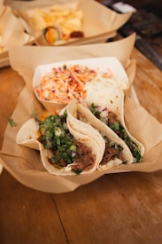 A rustic table holds a serving of street tacos, filled with seasoned meat topped with fresh cilantro and onions. Beside the tacos, a small dish contains a vibrant coleslaw with shredded carrots and cabbage. The entire meal is placed on a piece of brown parchment paper.