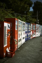 A row of various vending machines lined up along a street. There are multiple machines in different colors, including red, white, and blue. The area is surrounded by green foliage, and there is a crate at the base of the first vending machine.
