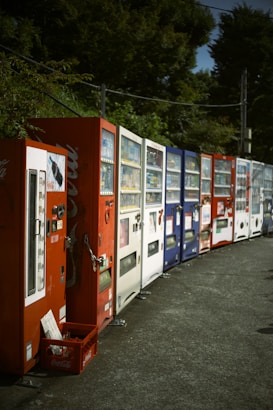 A row of various vending machines lined up along a street. There are multiple machines in different colors, including red, white, and blue. The area is surrounded by green foliage, and there is a crate at the base of the first vending machine.
