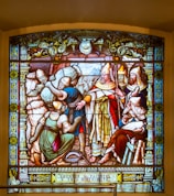 A detailed stained glass artwork depicting several figures, including a central clergy figure in ornate robes and a mitre, surrounded by attendants and townsfolk. The figures are engaged in a scene of distribution or charity, with a focus on the act of giving. The borders of the glass are richly decorated with intricate patterns and symbols.