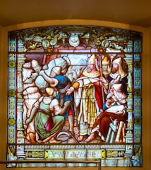 A detailed stained glass artwork depicting several figures, including a central clergy figure in ornate robes and a mitre, surrounded by attendants and townsfolk. The figures are engaged in a scene of distribution or charity, with a focus on the act of giving. The borders of the glass are richly decorated with intricate patterns and symbols.