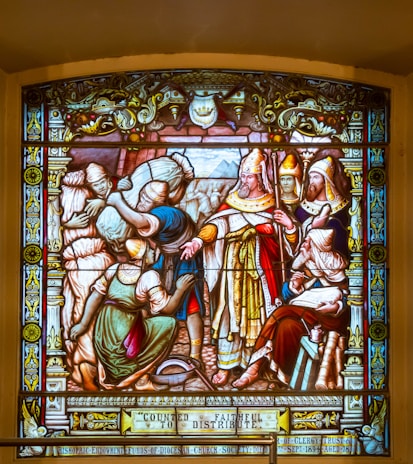 A detailed stained glass artwork depicting several figures, including a central clergy figure in ornate robes and a mitre, surrounded by attendants and townsfolk. The figures are engaged in a scene of distribution or charity, with a focus on the act of giving. The borders of the glass are richly decorated with intricate patterns and symbols.
