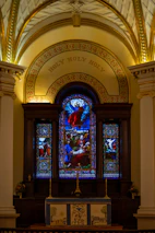 An ornate altar in a church featuring a stained glass window with religious imagery. The arched ceiling is adorned in gold accents, and the words 'Holy Holy Holy' are inscribed above. The altar includes elaborate decorations and floral arrangements.