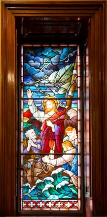 A stained glass window depicting a biblical scene on a turbulent sea. A central figure with a halo and raised hand stands beside a boat. Several people, some looking worried, are on the boat with a mix of expressions. The sky is filled with dark clouds and lightning, and the sea below is shown with waves crashing against the boat.