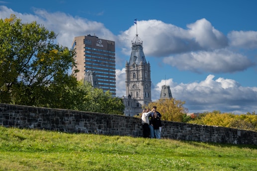 A group of people stand on a grassy hill in front of a stone wall, with a view of a cityscape consisting of a tall hotel building and a historic-looking government building with a flag on top. Trees with green and yellow leaves are visible, and the sky is bright blue with scattered clouds.