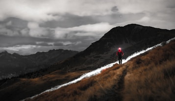 A lone hiker with a red jacket and headlamp walks along a grassy path on a hillside. The night sky looms overhead with clouds partially obscuring the moonlight, creating a dramatic atmosphere. The dark silhouette of mountains can be seen in the background.