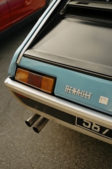 Close-up of the rear of a classic Renault car, with focus on its branding and taillight. The vehicle's exhaust pipes are visible beneath, and part of a red car is seen next to it.