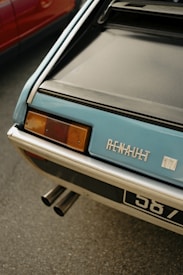 Close-up of the rear of a classic Renault car, with focus on its branding and taillight. The vehicle's exhaust pipes are visible beneath, and part of a red car is seen next to it.