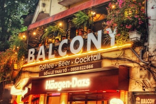 A lively café and bar space with vibrant signs and colorful illumination. The upper balcony is adorned with greenery and hanging lights, creating an inviting atmosphere. The signage prominently features the brands 'Balcôny' and 'Häagen-Dazs'. People are visible enjoying the ambiance on the balcony.